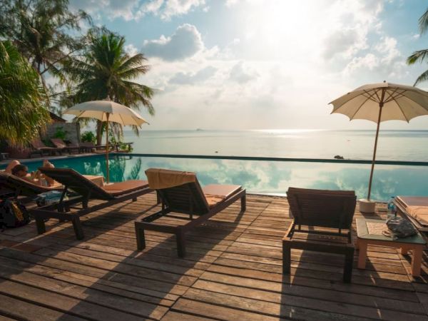 Deck chairs and umbrellas by a pool with a scenic ocean view, palm trees, and a clear sky in the background.