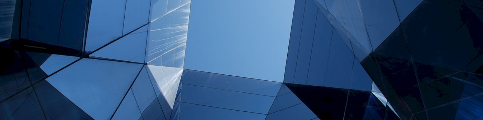 The image shows a geometric architectural structure with reflective glass panels, creating an angular pattern under a blue sky.