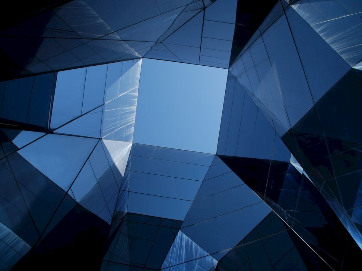 Geometric architecture with a central blue sky view, featuring reflective, angular glass panels creating a symmetrical, abstract scene.
