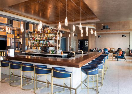 A modern bar with a wooden counter, blue stools, hanging lights, and shelves stocked with bottles, in a stylish open space.