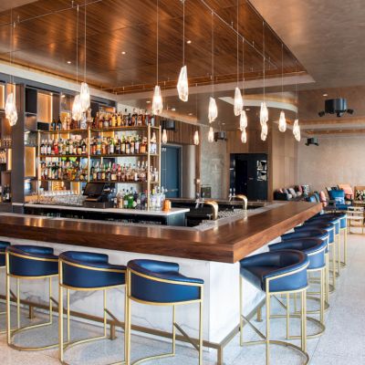 The image shows a modern bar with a large counter, bar stools, hanging lights, and shelves of bottles in an upscale setting.