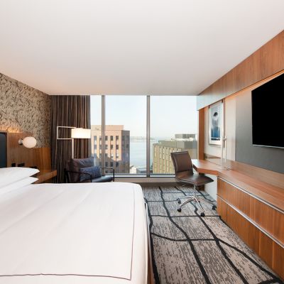 The image shows a modern hotel room with a large bed, wall-mounted TV, desk, chair, and a cityscape view from a large window.