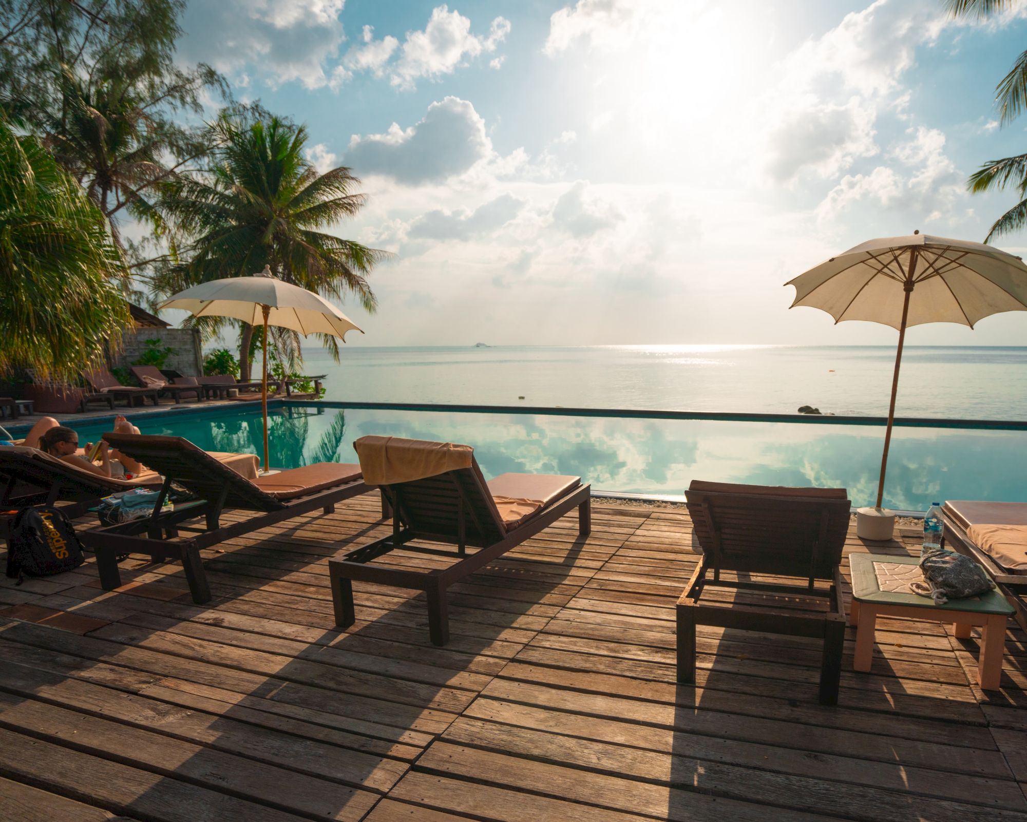A serene poolside scene with lounge chairs, umbrellas, and a view of the ocean under a bright, partly cloudy sky.