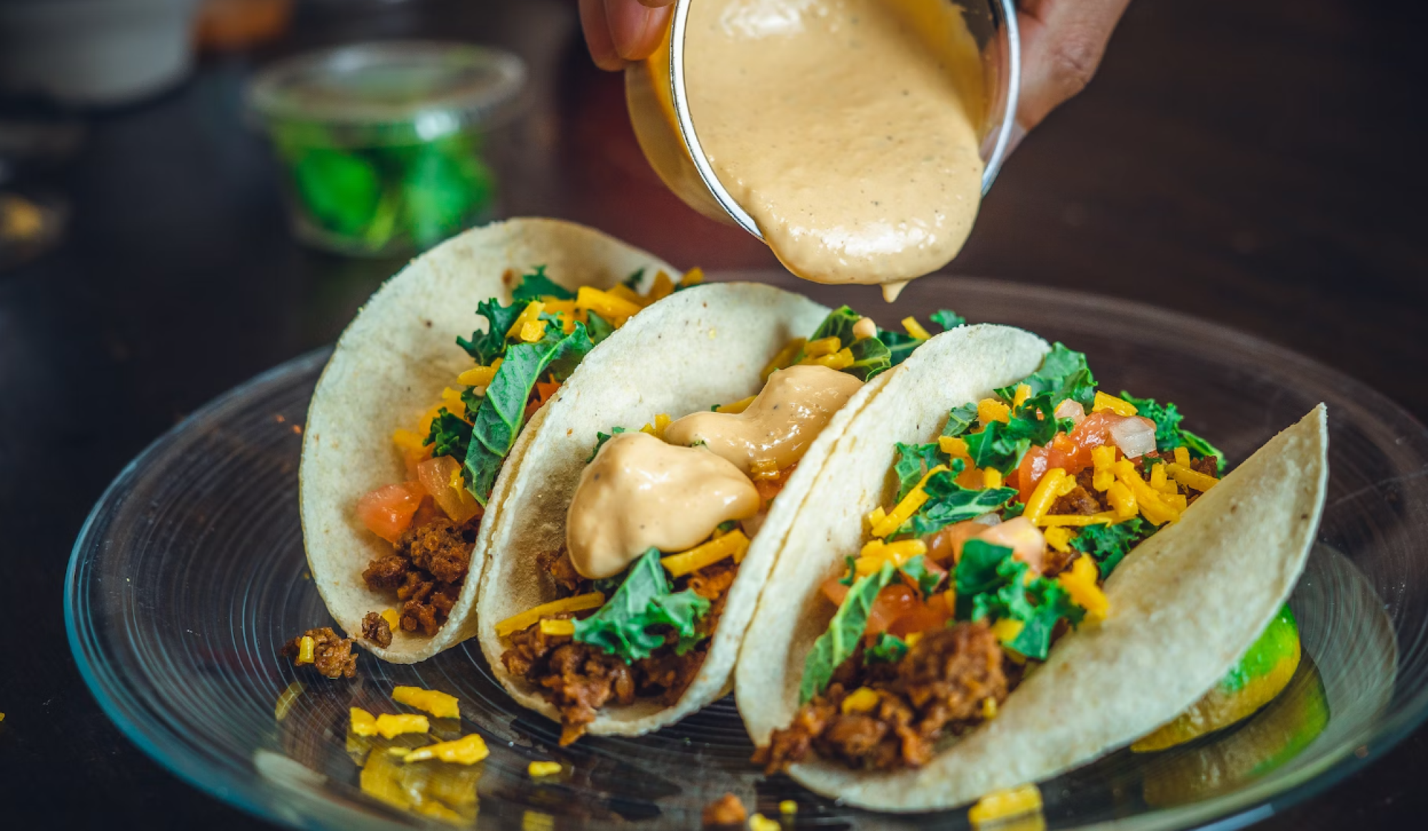Three tacos filled with seasoned meat, cheese, tomatoes, and greens are being topped with a creamy sauce from a small cup.
