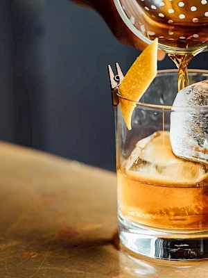 A cocktail is being poured into a glass with ice and an orange twist garnish on the rim.