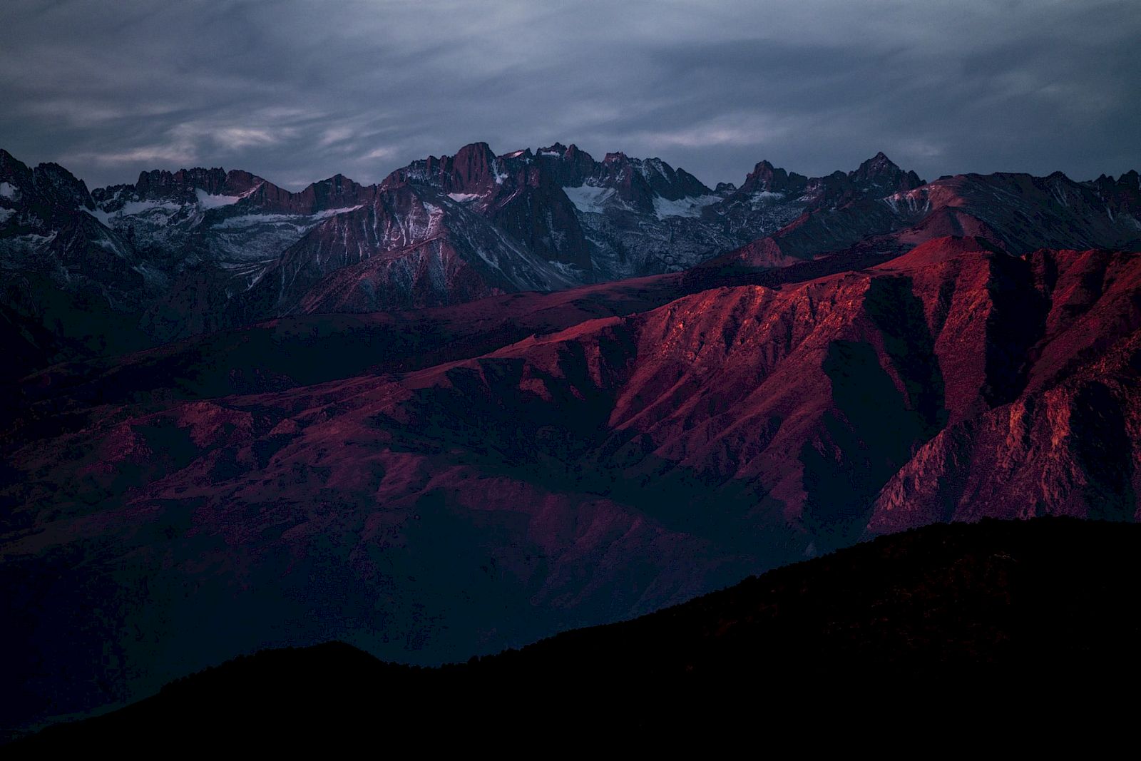 A range of mountains, tinted in deep red hues, under a cloudy sky, creating a dramatic and moody landscape scene.