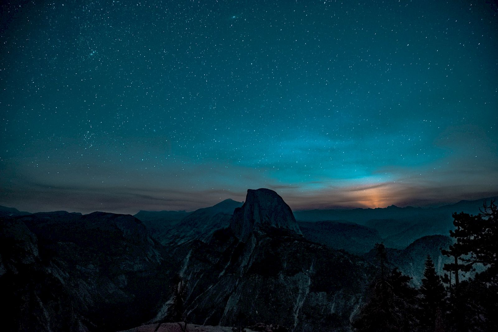 A starry night sky over silhouettes of mountains with a soft glow near the horizon in a tranquil landscape.