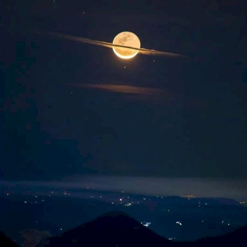 A full moon with clouds resembling Saturn's rings over a landscape with scattered lights at night.