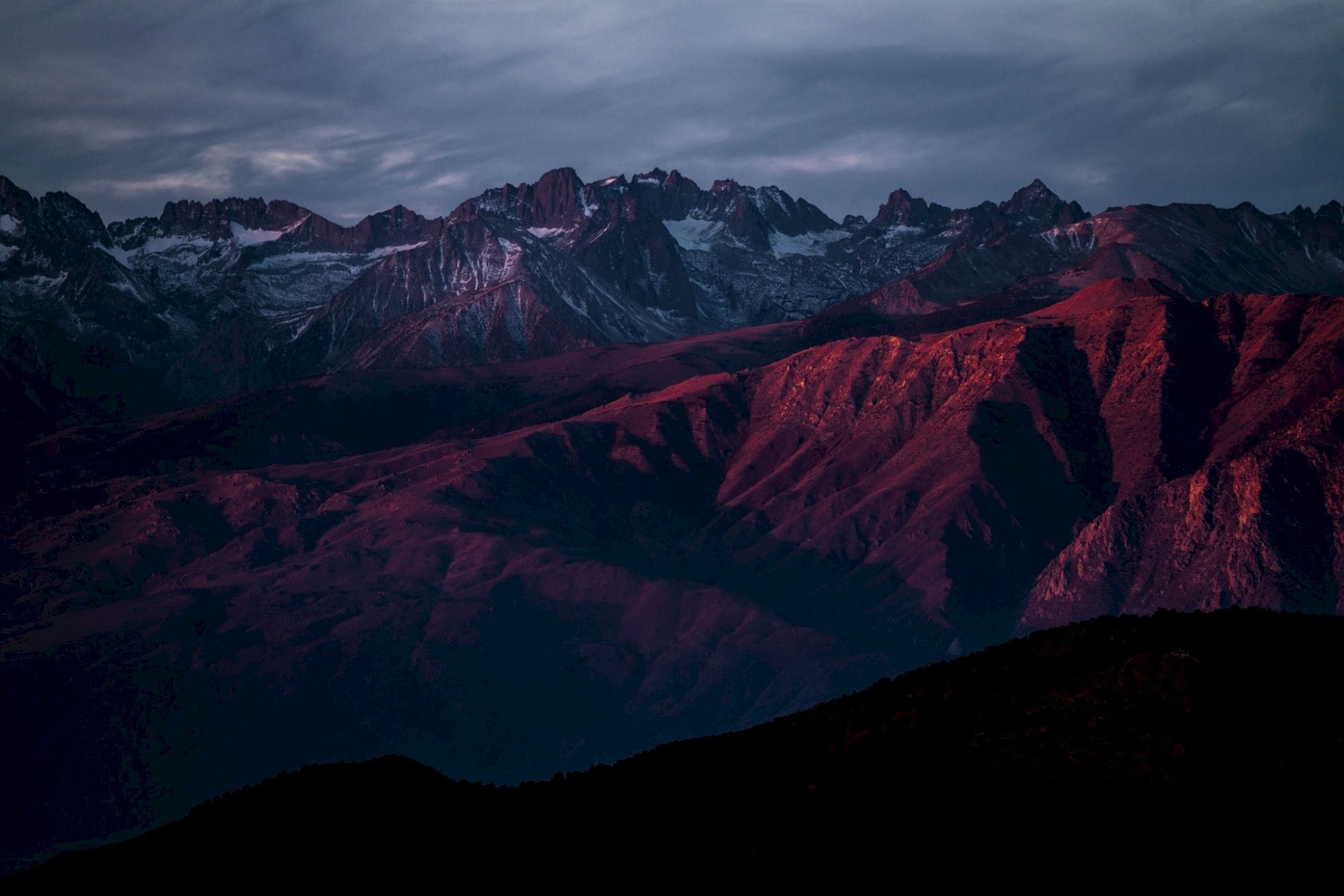 A mountain range illuminated by a reddish sunset under a cloudy sky.