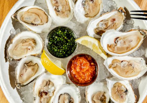 A plate of oysters on ice with lemon wedges and sauces, surrounded by two glasses, including one with an olive garnish on a pick.