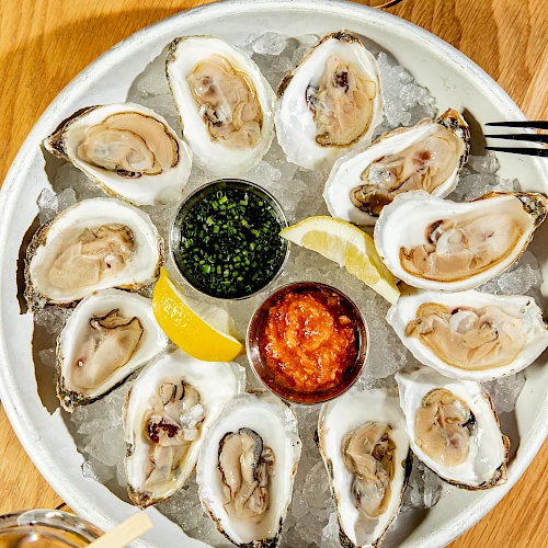 A plate of oysters on ice with lemon wedges and sauces, surrounded by two glasses, including one with an olive garnish on a pick.