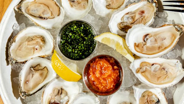 A plate of oysters on ice with lemon wedges and sauces, surrounded by two glasses, including one with an olive garnish on a pick.