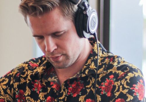 A person wearing headphones and a floral shirt is focused on something, with a plant and window in the background.