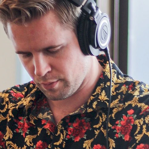 A person wearing headphones and a floral shirt is focused on something, with a plant and window in the background.