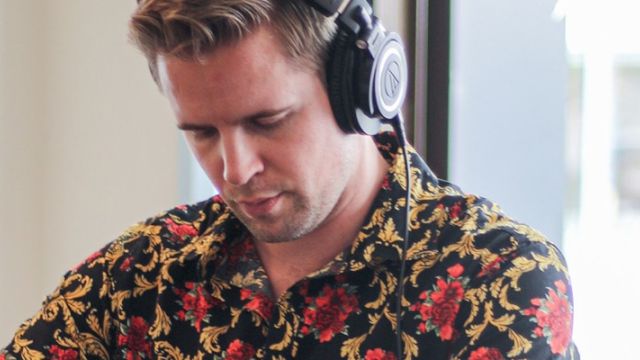 A person wearing headphones and a floral shirt is focused on something, with a plant and window in the background.
