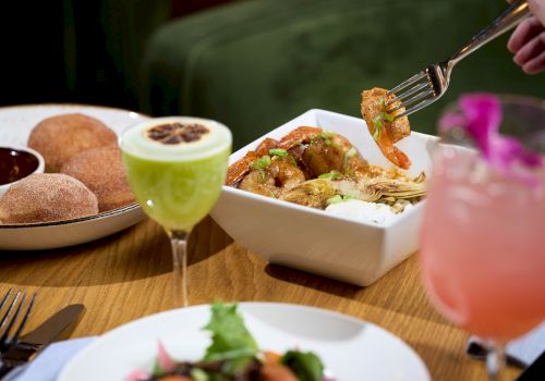 A meal with a dish of shrimp and vegetables, cinnamon sugar doughnuts, a green cocktail, a pink drink, and a salad on a dining table.