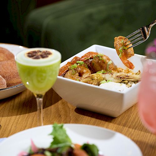 A meal with a dish of shrimp and vegetables, cinnamon sugar doughnuts, a green cocktail, a pink drink, and a salad on a dining table.