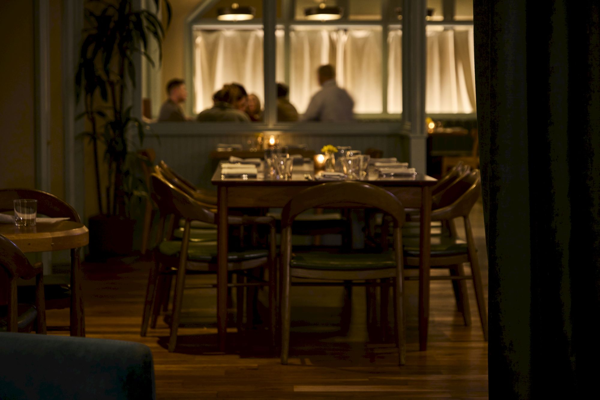 A dimly lit restaurant features an empty table in the foreground and a group of people conversing in the background behind glass panels.