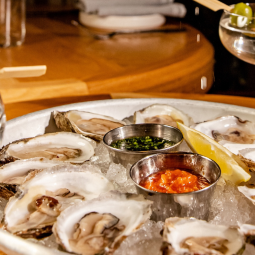 The image shows a plate of oysters on ice with lemon and sauces, accompanied by two martini glasses garnished with olives.