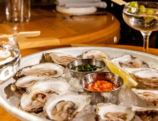 The image shows a plate of oysters on ice with lemon and sauces, accompanied by two martini glasses garnished with olives.