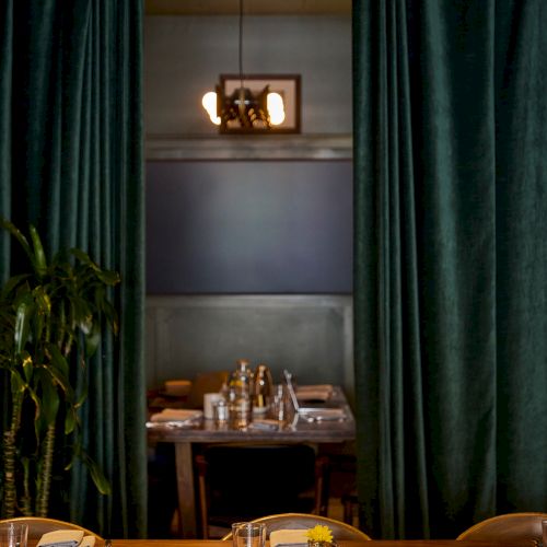 This image shows a dining area with a wooden table set for a meal, surrounded by green curtains and dim lighting for an intimate atmosphere.