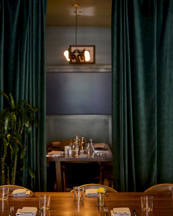This image shows a dining area with a wooden table set for a meal, surrounded by green curtains and dim lighting for an intimate atmosphere.