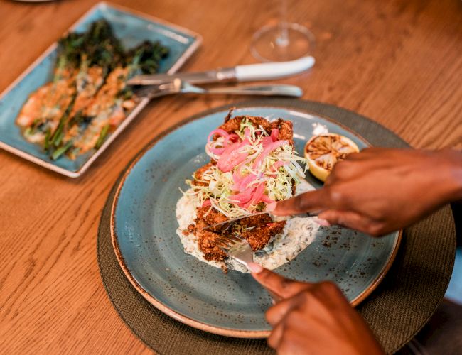 A person is preparing or eating vibrant tacos or flatbreads topped with veggies and sauce, with hands arranging toppings on a blue plate.