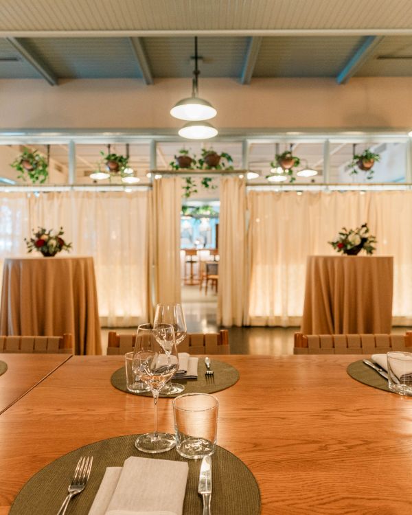 A cozy dining setup with a long wooden table, place settings, and soft window curtains; ambient lighting and potted plants decorate the space.