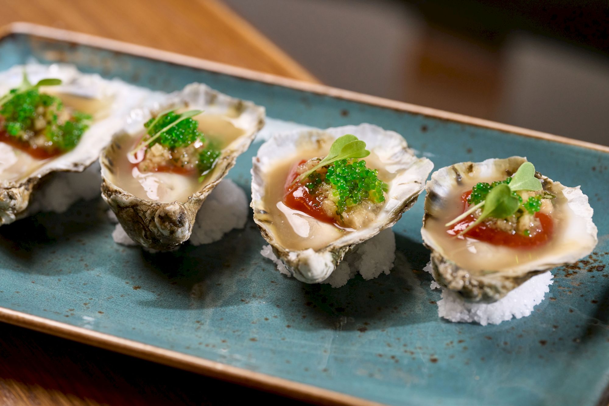This image shows a plate of gourmet oysters garnished with various toppings, served on a rectangular blue platter.