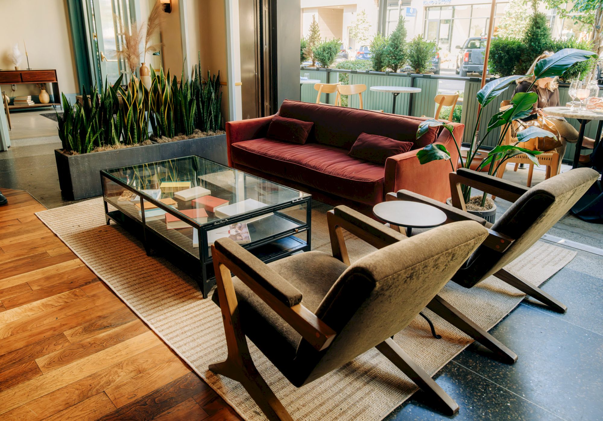 A cozy living room with a red sofa, two armchairs, a glass coffee table, plants, and large windows letting in natural light.