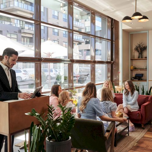 A group is seated at a table in a modern café, engaging in conversation, while a person at a podium works on a laptop.