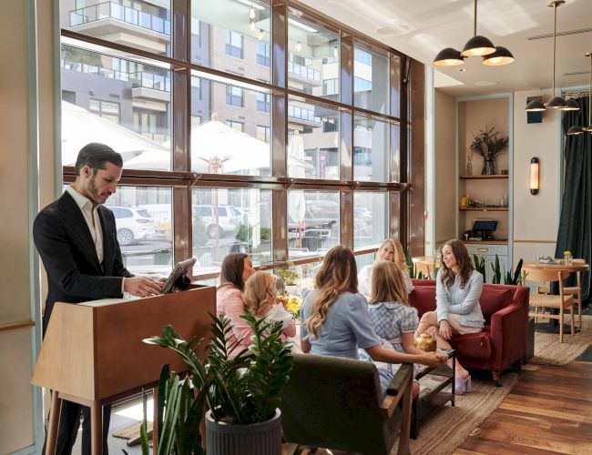 A group is seated at a table in a modern café, engaging in conversation, while a person at a podium works on a laptop.