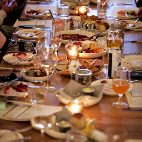 A long dining table filled with plates of food, glasses, and lit candles, surrounded by people eating and drinking.