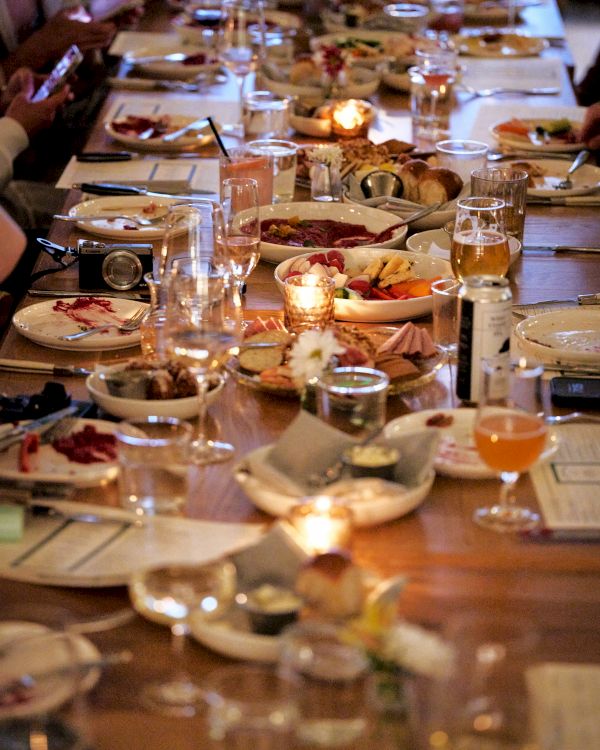 A long dining table filled with plates of food, glasses, and lit candles, surrounded by people eating and drinking.