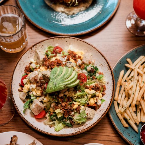 A colorful, crunchy bowl with greens, avocado, seeds, and diced toppings, surrounded by fries and drinks on a wooden table.