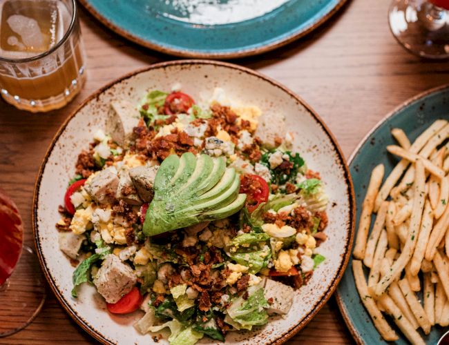 A colorful, crunchy bowl with greens, avocado, seeds, and diced toppings, surrounded by fries and drinks on a wooden table.