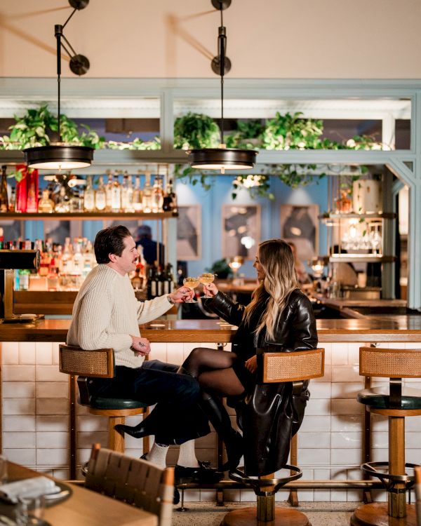 Two people toast drinks at a cozy bar with warm lighting, stools, and plants on a shelf behind the counter.