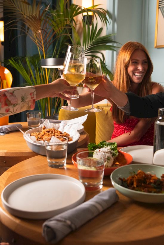 People dining together, clinking glasses, and smiling. Plates of food are on the table, creating a lively and cheerful atmosphere.
