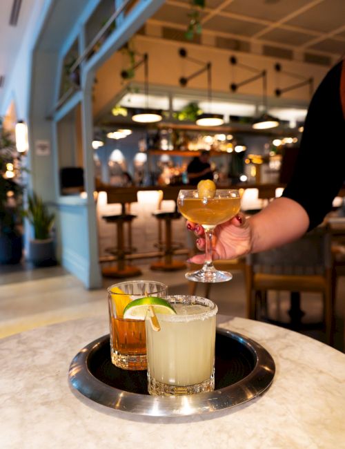 Three cocktails on a table in a bar setting, with someone lifting one glass on a tray.