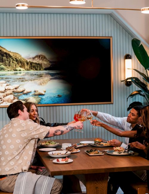 A group of four friends toasting with drinks at a wooden dining table, sharing a meal in a cozy, coastal-themed room with a landscape painting.