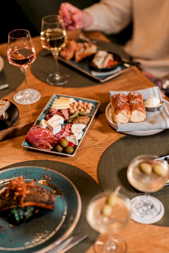 A shared dinner scene with tapas, small plates of meats and cheeses, pastries, and drinks on a wooden table.