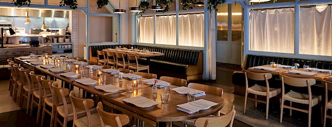 The image shows a warmly lit, modern restaurant interior with wooden tables, chairs, and hanging lights, giving a cozy ambiance.