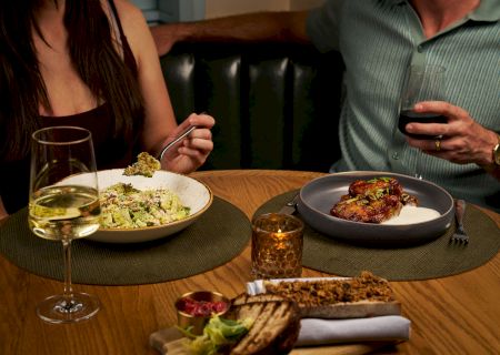 Two hands on a table with plates of food; a small finished dish on the left and a darker dish on the right, plus cutlery and a few items nearby. The scene suggests a meal setup.