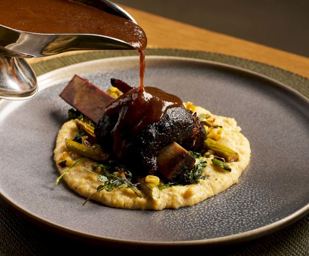 A plate with mashed potato or puree topped with rich red sauce and chunks of meat, drizzle pouring from a spoon above, plated artfully.