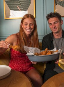 A couple is seated at a dining table, sharing fried food and smiling, with wine glasses and elegant decor in the background.