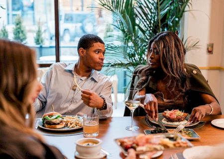 Two people are dining and chatting at a table with plates, drinks, and a shared appetizer, while a third person watches nearby.