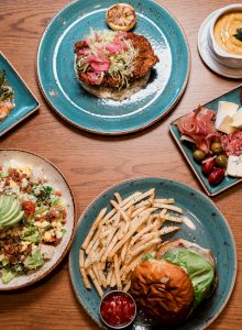A spread of colorful dishes: burgers, quesadillas, a salad with avocado, assorted appetizers, grilled meats, fries, and a small dessert&mdash;delicious plates all around.
