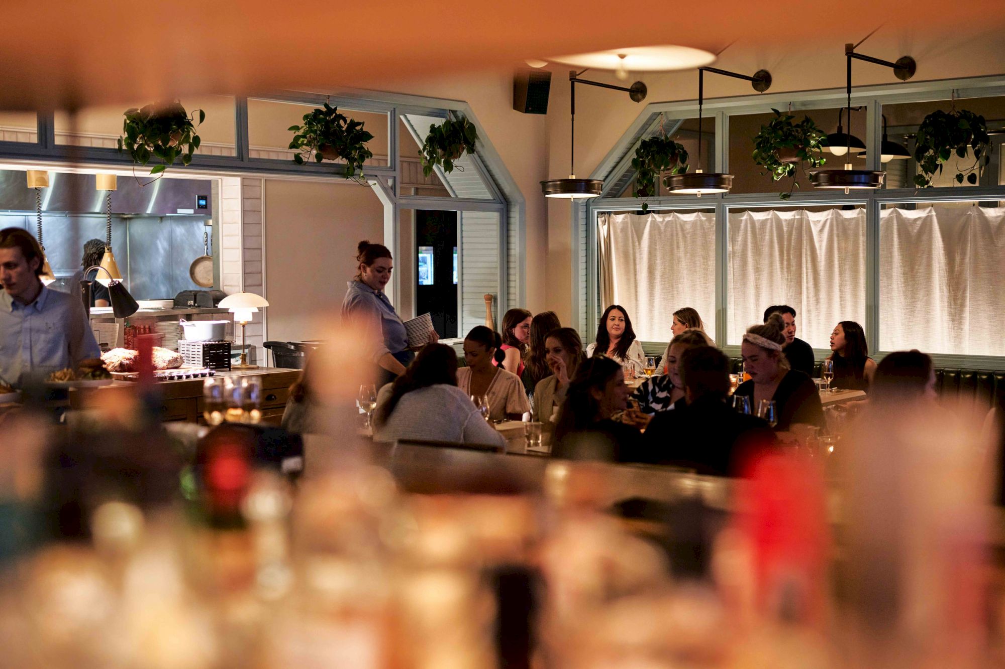 A lively restaurant scene with people dining, soft lighting, plants above windows, and a blurred foreground with bottles.