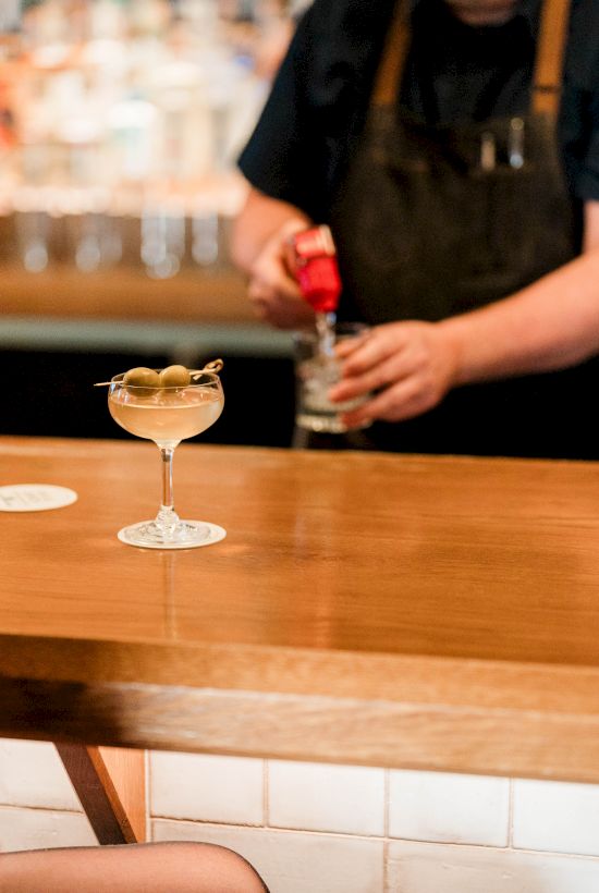 A bar scene with a bartender pouring a drink, two cocktails on the wooden counter, and a person sitting nearby, casual evening vibe.