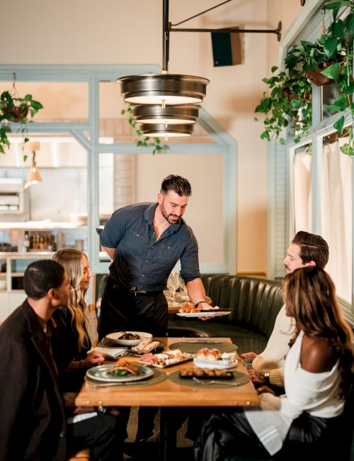 A waiter serves a plate to a group at a cozy restaurant, everyone chatting and smiling around a wooden table with shared dishes.